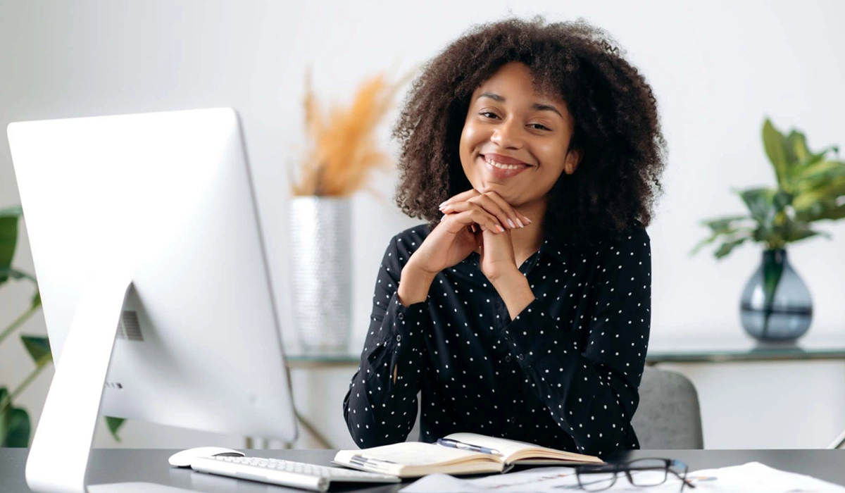 Foto de secretária empresarial sorridente, na mesa de trabalho em frente ao computador.